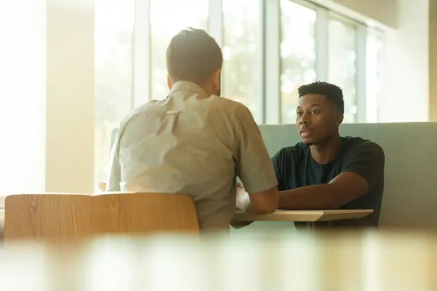 Two people talking across a table beside a window